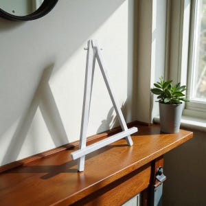 May include: A small, white wooden easel stands on a dark brown wooden surface. The easel is triangular, with a horizontal bar at the bottom. A small potted plant sits to the right of the easel. The background is a light-colored wall and a window.