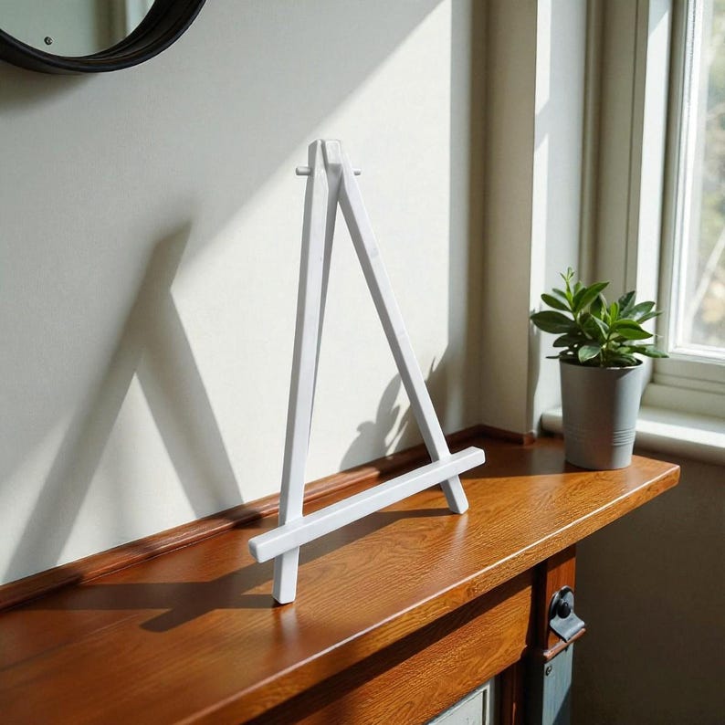 May include: A small, white wooden easel stands on a dark brown wooden surface. The easel is triangular, with a horizontal bar at the bottom. A small potted plant sits to the right of the easel. The background is a light-colored wall and a window.
