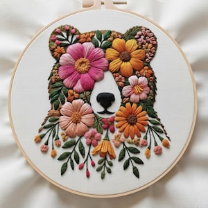 Op de afbeelding: Geborduurde bloemenberenkunst. De berenkop is gemaakt van kleurrijke bloemen in roze, oranje en gele tinten, met groene bladeren. Het gezicht van de beer is wit met een zwarte neus. De borduursels zitten in een houten hoepel.