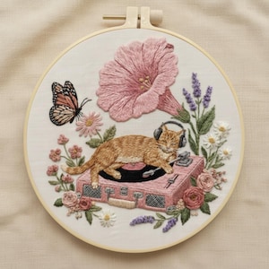 Op de afbeelding: Geborduurd kunstwerk in een houten hoepel. Het toont een oranje kat met een koptelefoon, liggend op een roze platenspeler. Een grote roze bloem, vlinder en andere bloemen omringen de kat. De borduursels zijn gedetailleerd.