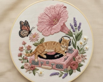 May include: Embroidered hoop art featuring a ginger cat wearing headphones, lounging on a pink record player. A large pink flower, butterfly, and various other flowers surround the cat. The embroidery is set in a wooden hoop.