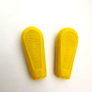 Puede incluir: Dos piezas alargadas de color amarillo brillante, de goma o plástico, con la parte superior redondeada. La palabra "PATENTED" está impresa verticalmente en cada pieza. La parte inferior de cada pieza tiene un borde ligeramente rugoso y texturizado. El texto "_ _ _ .RACEVELO. _ _ _" está en la parte inferior.