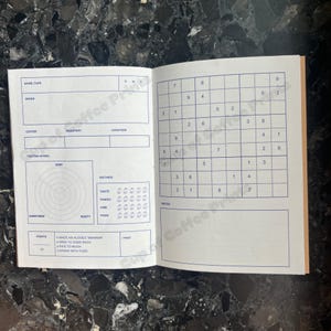 Puede incluir: Un cuaderno abierto con l&iacute;neas y texto impresos en azul sobre papel blanco. La p&aacute;gina izquierda presenta campos para detalles de caf&eacute;, una rueda de cata y valoraciones. La p&aacute;gina derecha contiene una cuadr&iacute;cula de Sudoku y notas.