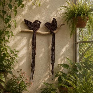 Puede incluir: Dos pájaros de macramé en color marrón oscuro hechos de cuerda anudada, con largas plumas de cola colgantes. Los pájaros están posados sobre un trozo de madera flotante de color claro, creando una pieza decorativa de pared con diseño texturizado.
