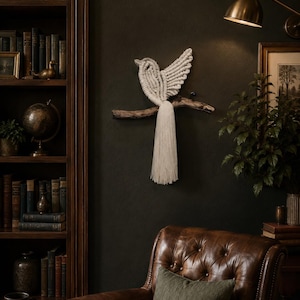 May include: A wall hanging featuring a cream-colored macrame bird crafted with intricate knotted cord patterns. The bird is perched on a natural, dark brown wooden branch, with a long, flowing fringe tail extending downward from the branch.