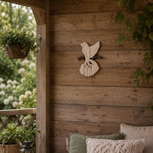 May include: A cream-colored macrame bird wall hanging crafted from knotted cord, featuring a detailed wing and tail design, mounted on a small dark brown wooden branch, displayed as a decorative piece on a rustic wooden wall surface.