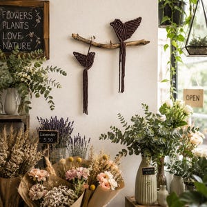 May include: Two dark brown macrame birds crafted from cord, featuring long fringed tails, are mounted on a natural wooden branch. The birds are displayed on a wall above various bouquets of dried flowers, lavender, and eucalyptus in a shop.