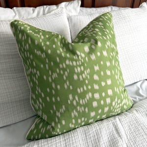 May include: A square decorative pillow with a green background and a pattern of small, irregular white shapes. The pillow has a white border and is placed on a white bed with other pillows.