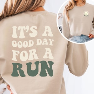 May include: A beige crewneck sweatshirt featuring the text IT'S A GOOD DAY FOR A RUN in large, wavy, cream and sage green lettering on the back. The front chest area displays a small circular graphic with a smiley face and the same text.