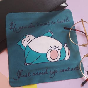 May include: Teal cleaning cloth featuring a cartoon Snorlax illustration with the text "If you don't want to battle, just avoid eye contact." The Snorlax is blue and white with pink accents. A black case and glasses are in the background.