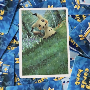 May include: A trading card featuring a Mimikyu character in a forest setting. The card has a white border and is surrounded by other blue cards with cartoon characters and text. The Mimikyu character is light yellow with black accents.