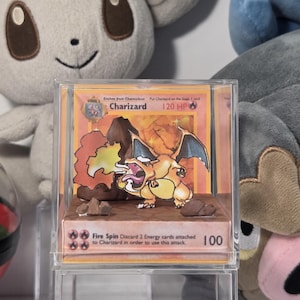 May include: A Charizard Pokémon card displayed in a clear acrylic case. The card features a fiery orange and yellow dragon with blue wings. The card text includes "Charizard", "120 HP", and "Fire Spin".