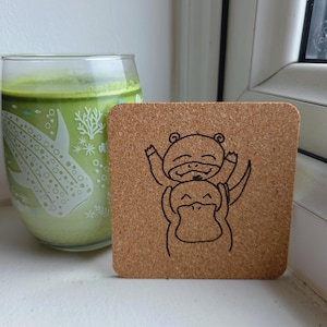 May include: A square cork drinks coaster featuring a black line drawing of two cartoon animals embracing. A glass of green drink is to the left of the coaster. The glass has a white design of sea creatures and plants.