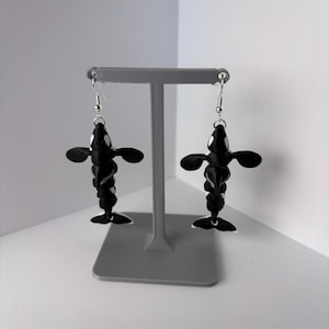 May include: A pair of dangling earrings featuring articulated orca whale charms. Each charm is black with white eye patches and a white belly, suspended from silver-toned metal fishhook wires. The orca figures have distinct fins and tails.
