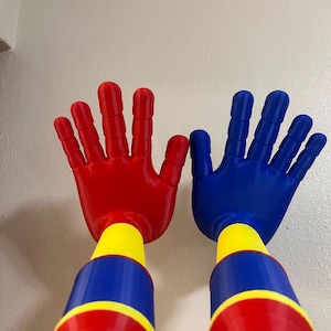 Könnte beinhalten: Zwei farbenfrohe, handförmige Spielzeuge. Eines ist rot, das andere blau. Jedes hat einen gelben und blauen Griff. Die Spielzeuge sind so gestaltet, dass sie großen Händen ähneln, mit ausgestreckten Fingern. Die Spielzeuge sind aus Kunststoff.