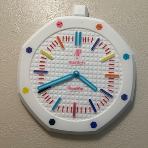 Könnte beinhalten: Weiße achteckige Wanduhr mit einem gitterförmigen Zifferblatt. Die Uhr hat farbige Stundenmarkierungen und Zeiger mit dem Text "AP x Swatch" und "Royal Pop". Sie hat ein kleines weißes rechteckiges Stück oben.