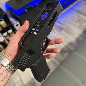 May include: A black plastic remote control holder shaped like a handgun. The holder features a cutout to securely fit a Roku remote with a purple directional pad, volume buttons, and shortcut buttons for Netflix, Disney Plus, and Roku Channel.