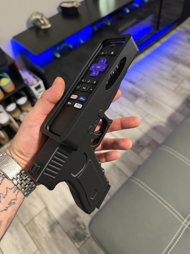 May include: A black plastic remote control holder shaped like a handgun. The holder features a cutout to securely fit a Roku remote with a purple directional pad, volume buttons, and shortcut buttons for Netflix, Disney Plus, and Roku Channel.