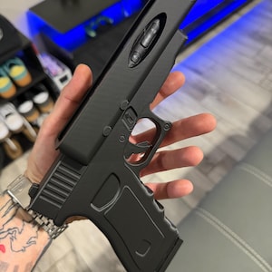 May include: A matte black handheld device shaped like a firearm, featuring a top-mounted control panel with three circular buttons. The object has a textured grip, a trigger guard, and a sleek, solid-coloured frame with a distinct rectangular top section.