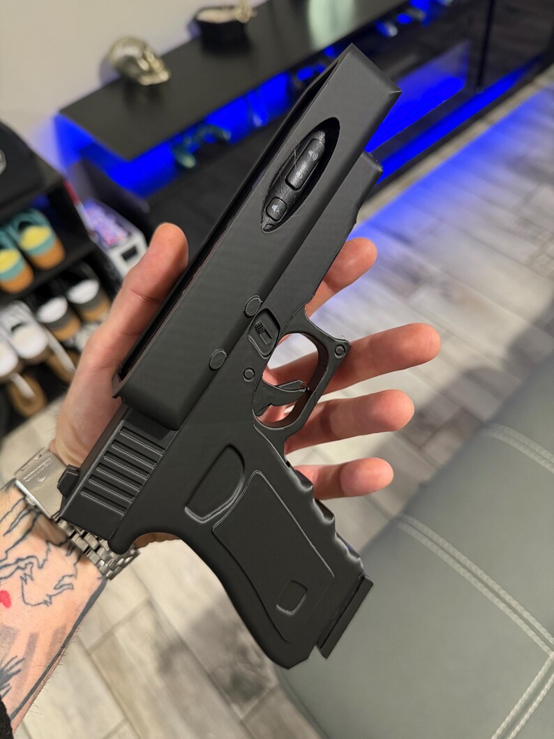 May include: A matte black handheld device shaped like a firearm, featuring a top-mounted control panel with three circular buttons. The object has a textured grip, a trigger guard, and a sleek, solid-coloured frame with a distinct rectangular top section.