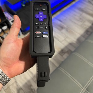 May include: A black remote control encased in a protective black silicone sleeve. The remote features a purple directional pad with an OK button, volume controls, and dedicated buttons for Netflix, Disney Plus, Apple TV Plus, and the Roku Channel.