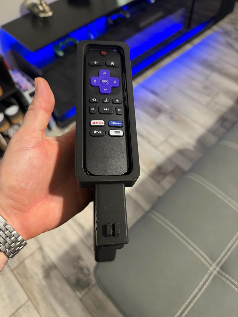 May include: A black remote control encased in a protective black silicone sleeve. The remote features a purple directional pad with an OK button, volume controls, and dedicated buttons for Netflix, Disney Plus, Apple TV Plus, and the Roku Channel.