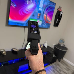May include: A black plastic remote control holder shaped like a firearm, featuring a purple directional pad and dedicated streaming service buttons. The device is held upright, pointing towards a large television screen displaying a colourful digital interface.