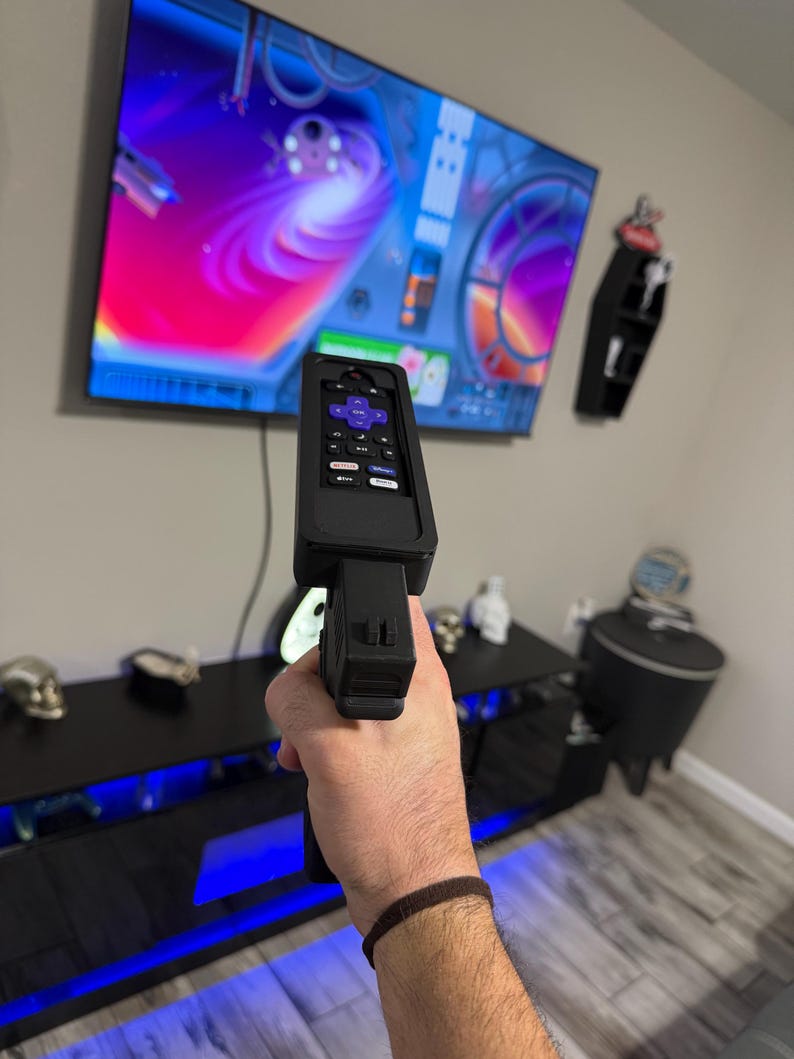 May include: A black plastic remote control holder shaped like a firearm, featuring a purple directional pad and dedicated streaming service buttons. The device is held upright, pointing towards a large television screen displaying a colourful digital interface.