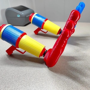 May include: Two brightly coloured water pistols. The water pistols have red handles, yellow barrels, and blue and red nozzles. One is angled to the right. The other is in the background.