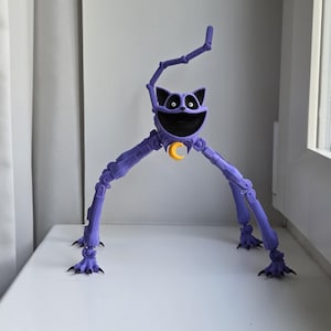 May include: A purple articulated cat figurine with long, jointed legs and a wide, black-lined mouth. The figure features a yellow crescent moon charm hanging from its neck, large white eyes with black pupils, and sharp black claws on its feet.