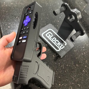 May include: A black plastic remote control holder shaped like a handgun, featuring a purple directional pad and various media buttons. Beside it sits a matching black display stand with the text GLOCKU printed in white on its rectangular base.