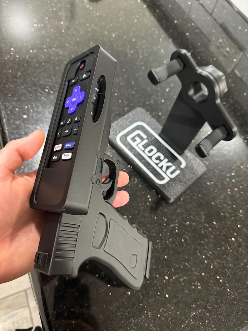 May include: A black plastic remote control holder shaped like a handgun, featuring a purple directional pad and various media buttons. Beside it sits a matching black display stand with the text GLOCKU printed in white on its rectangular base.