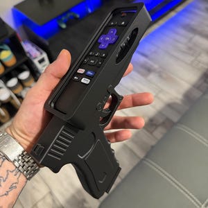 May include: A black plastic remote control holder shaped like a handgun. The holder features a cutout to accommodate a Roku remote with a purple directional pad and buttons for Netflix, Disney Plus, and The Roku Channel, all set within a matte frame.