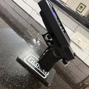 May include: A black remote control holder shaped like a handgun, featuring a textured grip and trigger guard. The holder is mounted on a rectangular black base with the white text GLOCK printed on the top surface, designed to hold a television remote.