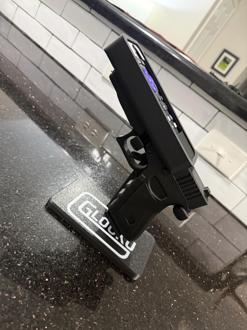 May include: A black remote control holder shaped like a handgun, featuring a textured grip and trigger guard. The holder is mounted on a rectangular black base with the white text GLOCK printed on the top surface, designed to hold a television remote.