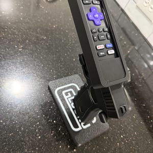 May include: A black plastic remote control holder shaped like a firearm, featuring a purple directional pad and various streaming service buttons. The holder is mounted on a rectangular black base with the white text GLOCK printed on the top surface.