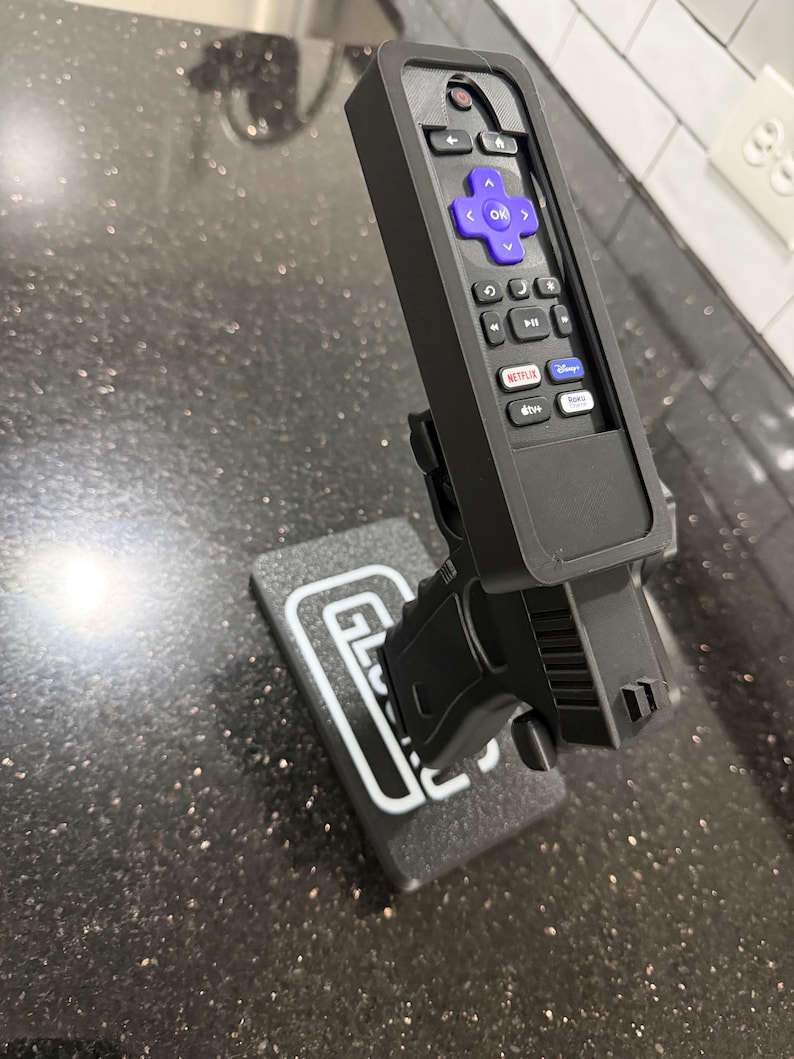 May include: A black plastic remote control holder shaped like a firearm, featuring a purple directional pad and various streaming service buttons. The holder is mounted on a rectangular black base with the white text GLOCK printed on the top surface.