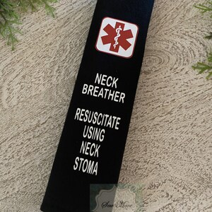 Op de afbeelding: Een zwarte nekbreather met een wit medisch symbool en de tekst "Neck Breather Resuscitate Using Neck Stoma".