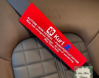 May include: Red car seat belt cover with white text that reads "AUTISM / NON-VERBAL - PANHYPOPITUITARYISM May not recognize danger. At risk of wandering. May resist help." and a blue medical symbol. The text "Kurt" is also on the cover in blue with a blue puzzle piece.