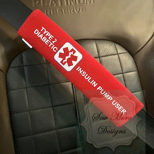 Puede incluir: Funda roja para cinturón de seguridad con el texto "Type 2 Diabetic Insulin Pump User" y un símbolo médico rojo con una serpiente enroscada alrededor de un bastón. El texto "Sew More Designs" es visible en la esquina inferior derecha.