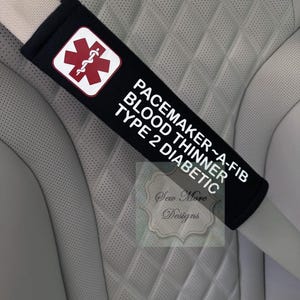 Puede incluir: Funda negra para cinturón de seguridad con texto blanco: PACEMAKER~A-FIB, BLOOD THINNER, TYPE 2 DIABETIC. Presenta un símbolo médico rojo sobre fondo blanco. La funda está sobre un cinturón blanco, sobre un asiento de cuero gris acolchado.