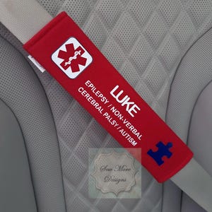 May include: A red seatbelt cover with white text that reads "LUKE" and "EPILEPSY / NON-VERBAL CEREBRAL PALSY / AUTISM." It features a white medical symbol and a blue puzzle piece. The cover is placed over a gray seatbelt.