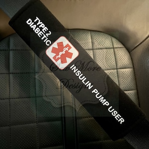 Puede incluir: Funda de cinturón de seguridad negra con texto blanco que dice "Tipo 2 Diabético Usuario de Bomba de Insulina". Un símbolo médico rojo con una serpiente está en el centro de la funda.