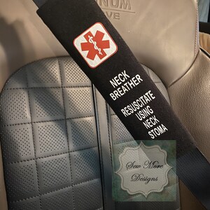 Op de afbeelding: Zwarte gordelhoes met een rood medisch symbool en de tekst "Neck breather resuscitate using neck stoma". De hoes is bevestigd aan een bruine leren autostoel. De tekst "Sew More Designs" is zichtbaar in de rechterbenedenhoek.