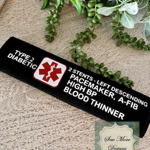 Puede incluir: Pulsera de alerta médica negra con texto blanco que dice "Tipo 2 Diabético, 2 Stents - Descendente Izquierdo Marcapasos, Fibrilación Auricular, Presión Arterial Alta, Diluyente de Sangre". Un símbolo médico rojo está en el lado izquierdo de la banda.
