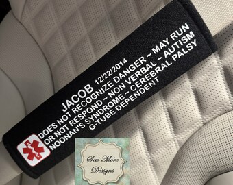 May include: Black car seatbelt cover with white text that reads "JACOB 12/22/2014 DOES NOT RECOGNIZE DANGER - MAY RUN OR NOT RESPOND - NON VERBAL - AUTISM NOONAN'S SYNDROME - CEREBRAL PALSY G-TUBE DEPENDENT".