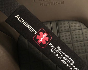 May include: Black seatbelt strap with a red medical symbol and the text "ALZHEIMERS" and "May not recognize danger. May resist help. At risk of wandering."  Sew More Designs is printed in a white circle.