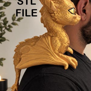 May include: A gold-colored dragon figurine with textured scales and large yellow eyes, featuring the text STL FILE printed in black. The dragon is positioned to rest on a shoulder, displaying detailed wings, a long tail, and a pointed head structure.