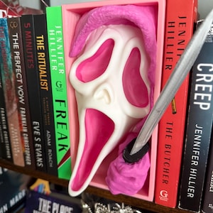 May include: A white and pink Scream mask is displayed in a pink box on a bookshelf. A silver knife is placed near the mask. Books with titles like "Freak" and "Creep" are visible.