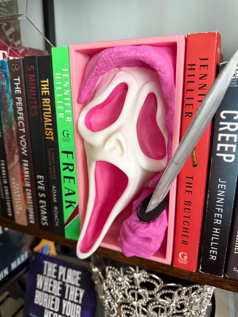 May include: A white and pink Scream mask is displayed in a pink box on a bookshelf. A silver knife is placed near the mask. Books with titles like "Freak" and "Creep" are visible.